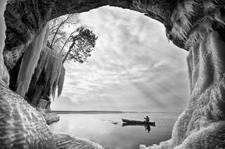 a canoeist glides on still waters as viewed through an ice cave in january