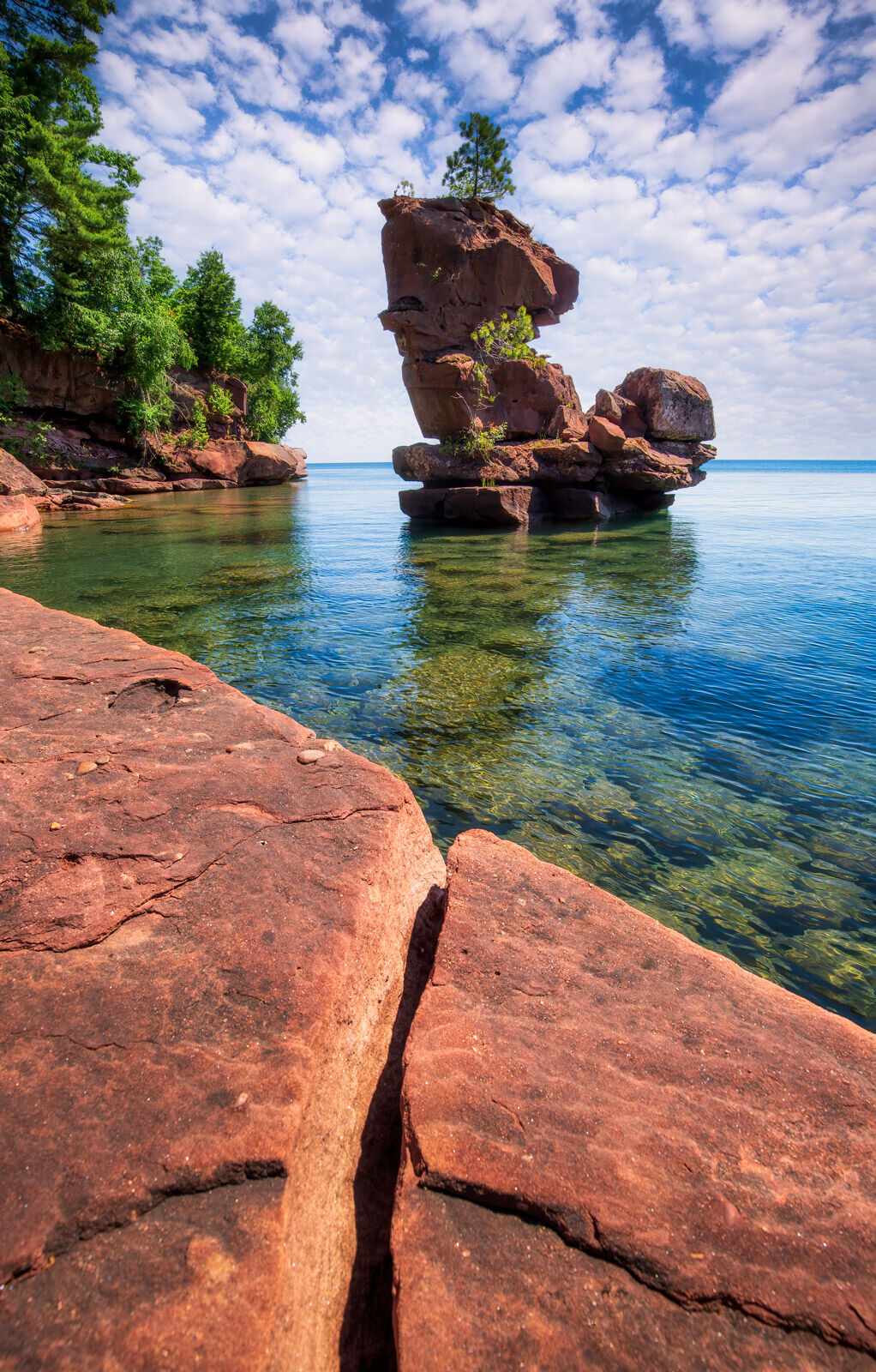 a sandstone sea stack appears to float on crystal clear water with puffy clouds in the sky